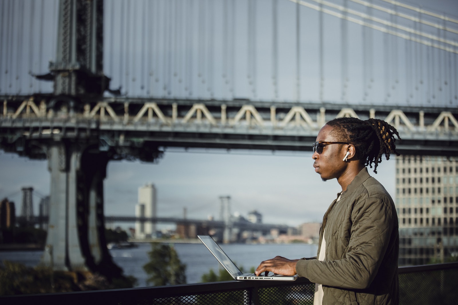 Side view of young African American male in sunglasses with dreadlocks and wireless earphones working on laptop placed on terrace railing against blurred Manhattan Bridge
